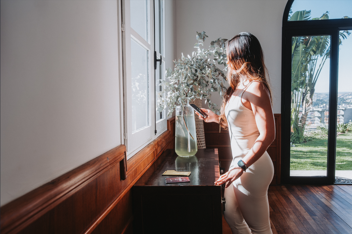 Femme regardant son téléphone près d'une fenêtre lumineuse, intérieur chaleureux avec vue sur jardin.