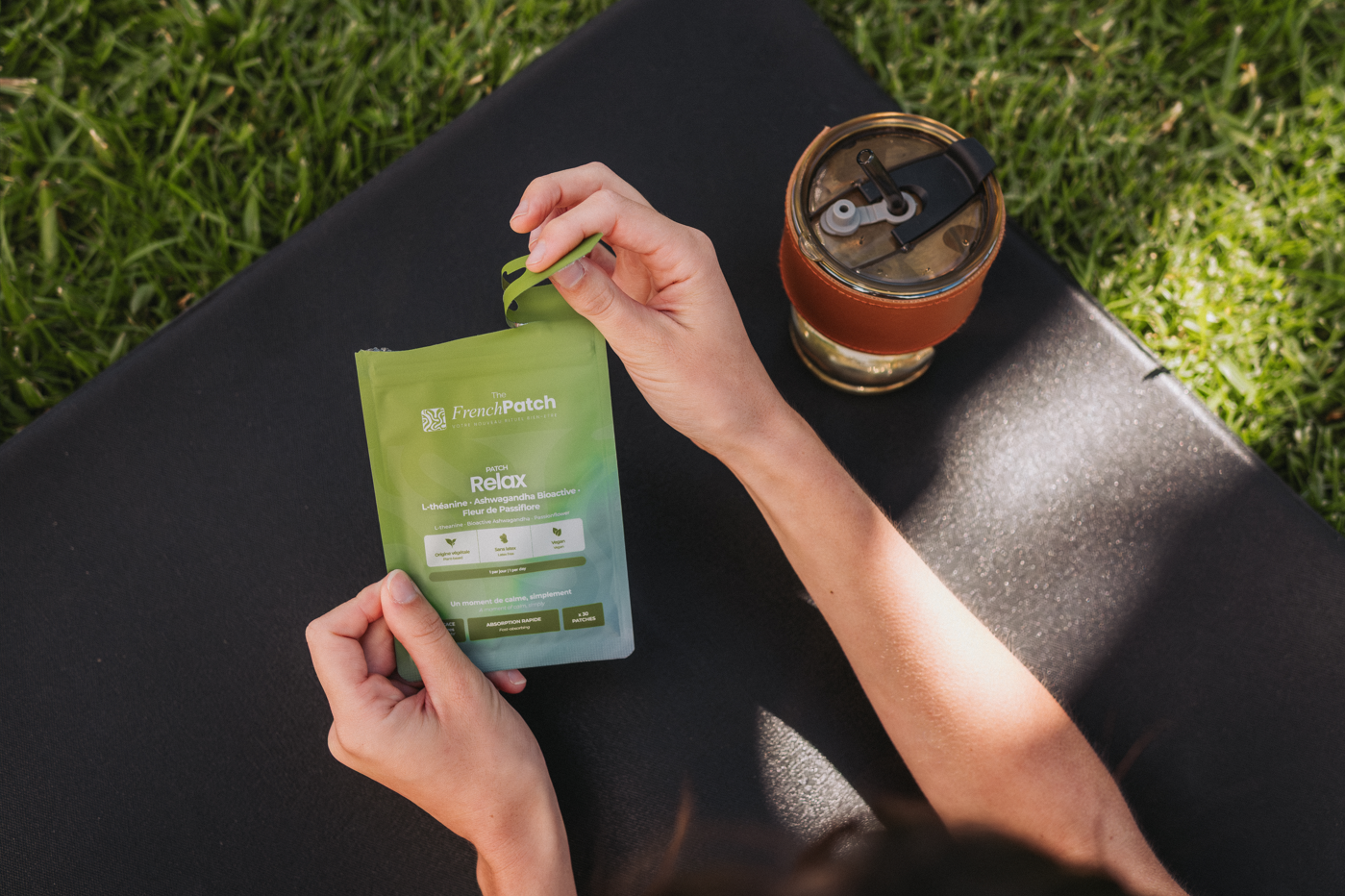 Femme ouvrant un sachet patch Relax The French Patch sur un tapis de yoga noir, avec une gourde isotherme à côté, en extérieur.